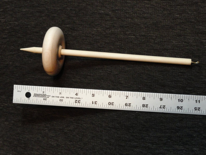 May include: A wooden drop spindle, a tool for hand spinning yarn. The spindle has a rounded whorl and a long, slender shaft. The shaft is approximately 10 inches long. The whorl is about 1.5 inches in diameter. The spindle is made of light-colored wood.