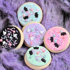 May include: Five decorated sugar cookies with different colored frosting and sprinkles. The cookies are decorated with bats and are on a purple fuzzy surface.