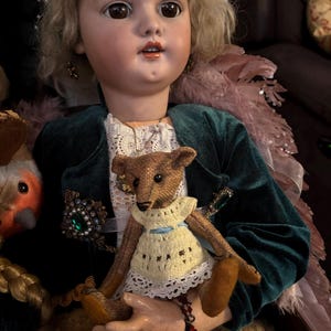 May include: A vintage porcelain doll with blonde hair and a green velvet jacket, holding a small teddy bear wearing a yellow dress. The doll has a pearl headpiece and a jeweled brooch.