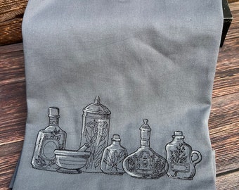 May include: A gray kitchen towel with an embroidered design of five glass bottles and a mortar and pestle. The bottles are filled with herbs and flowers.