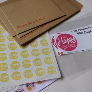 May include: A sheet of yellow and white round stickers with the text "The Happy Little Print Shop."  The stickers are packaged in a clear plastic envelope with a pink and white sticker that says "With Compliments."  The package is on top of a brown envelope with the text "Please do not bend" written in red.