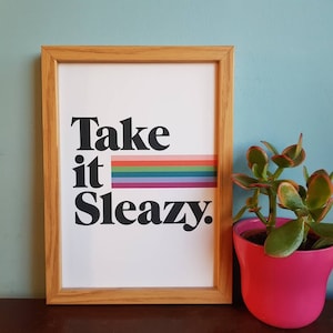 Könnte beinhalten: Ein gerahmter Druck mit dem Text "Take it Sleazy." in schwarzer Fettschrift. Ein Regenbogenstreifen befindet sich hinter dem Wort "it". Der Rahmen ist aus hellem braunem Holz.