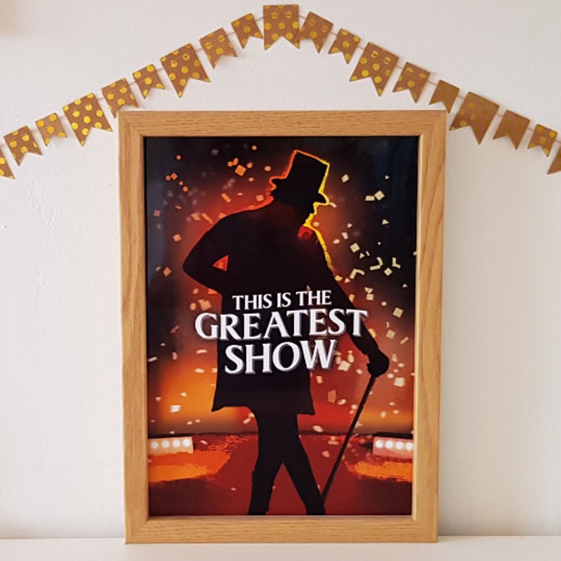 May include: A framed poster with a silhouette of a man in a top hat and tails. The poster has a red and orange background with confetti falling. The text "This is the Greatest Show" is printed in white on the poster.