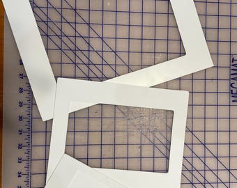 May include: Three white picture frames of different sizes, with a white mount board inside each frame, are arranged on a blue and white grid cutting mat.