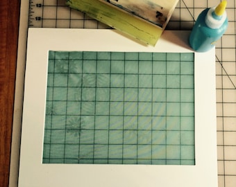 May include: A white mat with a green gridded screen inside. The screen is slightly translucent and shows a faint pattern. A blue squeegee and a bottle of blue liquid are visible in the background.