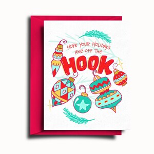 May include: A white card with a red envelope. The card has a festive design with colorful ornaments and the text "Hope your holidays are off the hook".