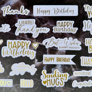 May include: A selection of white stickers with gold foil lettering. The stickers display phrases such as "Happy Birthday," "Thanks," "Hello," and "Best Wishes." Some stickers include heart-shaped accents. The background is a dark, marbled surface.
