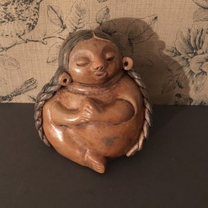 May include: A brown ceramic figurine of a woman with long braided hair. The woman is sitting with her hands clasped in front of her.