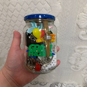 Puede incluir: Un frasco de vidrio con tapa azul lleno de varios abalorios, incluyendo un globo amarillo, un corazón rojo, una llave dorada y un letrero verde y negro que dice "GRAD".