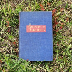 Puede incluir: Un libro de tapa dura azul con el título "A Treasury of Laughter" en letras doradas en la portada.