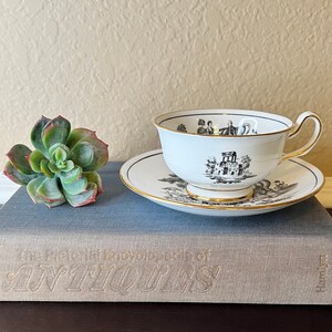 May include: A white teacup and saucer with black and gold trim. The teacup has a black and white design of a building and trees. The saucer has a black and white design of birds and flowers. The teacup and saucer are sitting on a gray book with the title "The Pictorial Encyclopedia of Antiques".
