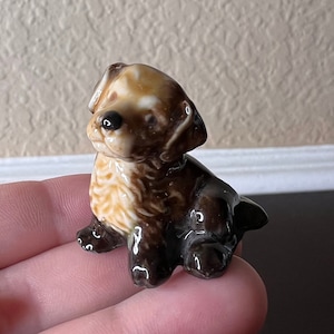 May include: A small ceramic figurine of a brown and white puppy sitting with its head tilted to the side. The puppy has a brown nose and black eyes.