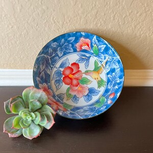 May include: A blue and white ceramic bowl with a floral and hummingbird design. The bowl has a blue rim and a white interior. The floral design features red, pink, and yellow flowers. The hummingbird is flying over the flowers and is yellow, orange, and red.
