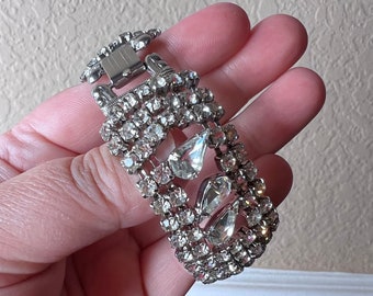 May include: A silver bracelet with a row of clear rhinestones and a large, clear, teardrop-shaped rhinestone in the center.