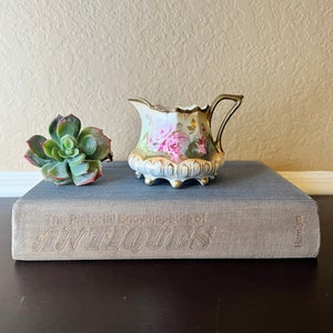 May include: A small, white porcelain creamer with a gold trim and floral design. The creamer has a handle and a scalloped rim. It is sitting on top of a book titled "The Pictorial Encyclopedia of Antiques".