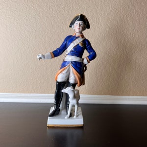 May include: A porcelain figurine of a man in a blue and white military uniform with a black hat, standing with a white dog at his side. The figurine is on a white base with gold trim.