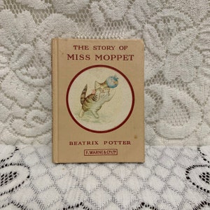 May include: A vintage copy of "The Story of Miss Moppet" by Beatrix Potter. The book features a tan cover with the title and author's name in red and black text. A circular illustration shows a playful tabby cat. The book is from F. Warne & Co Ltd.