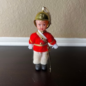 May include: A vintage plastic doll ornament dressed as a soldier. The doll is wearing a red uniform with gold trim, a white sash, and a gold helmet with a crest. The doll is holding a sword and has a white plume on its helmet.