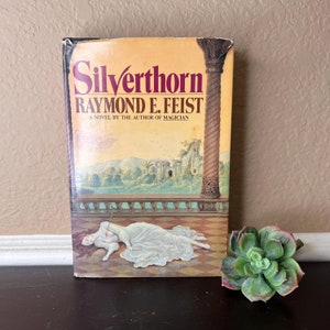 Pode incluir: Um livro de bolso vintage intitulado "Silverthorn" de Raymond E. Feist. A capa apresenta uma mulher com um vestido branco em um chão quadriculado, com uma cena de castelo. Uma planta suculenta verde está ao lado do livro.