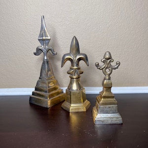 May include: Three decorative metal finials in gold and silver tones. The finials are shaped like fleur-de-lis and have a pointed top. They are all on a wooden surface.