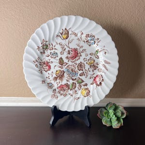 以下が含まれることがあります： 波打つ縁取りと、茶色、ピンク、黄色、青、緑の色合いの花柄が施された白いセラミック製のディナー皿です。