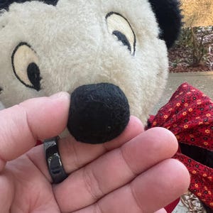 May include: A black ring is held in front of a plush toy's black nose. The toy has a white face with black ears and eyes. A red bow with a floral pattern is visible.