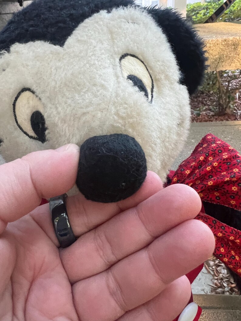 May include: A black ring is held in front of a plush toy's black nose. The toy has a white face with black ears and eyes. A red bow with a floral pattern is visible.