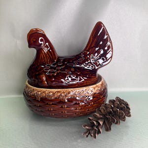May include: A brown ceramic hen-shaped dish with a basket weave base. The hen has a glossy finish and is perched atop the basket. A pine cone rests beside the dish. This decorative piece is suitable for kitchen decor.