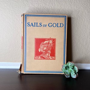 Puede incluir: Un libro antiguo de tapa dura con un borde azul y blanco y el título "SAILS OF GOLD" impreso en negro. La portada presenta una ilustración roja de un barco con velas.