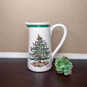 Op de afbeelding: Witte keramische kan met een groene rand en handvat. De kan heeft een kerstboomontwerp met ornamenten, cadeaus en de kerstman. Het woord "Spode" staat op de kan.