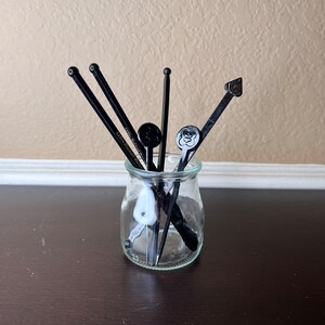 May include: A clear glass jar with a black lid filled with black and white cocktail stirrers. The stirrers have different designs and are arranged vertically in the jar.