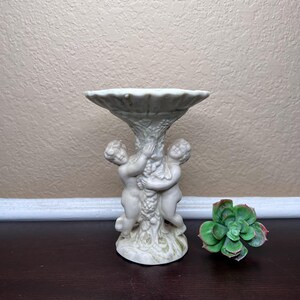 Puede incluir: Una fuente de pedestal de cerámica blanca con una parte superior acanalada, sostenida por dos figuras de querubines. Las figuras abrazan una columna central adornada con racimos de uvas. Una planta suculenta verde se encuentra a la derecha de la fuente.