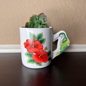 May include: White ceramic mug with a hummingbird handle and red hibiscus flower design. The mug is filled with a green succulent plant.