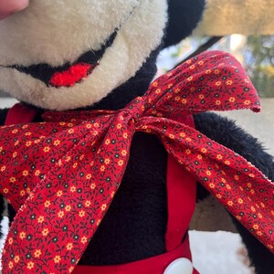 May include: A vintage plush toy featuring a black and white face with a red smile. The toy wears a red bow tie with a floral pattern and red overalls with a white button. The toy is a classic character.
