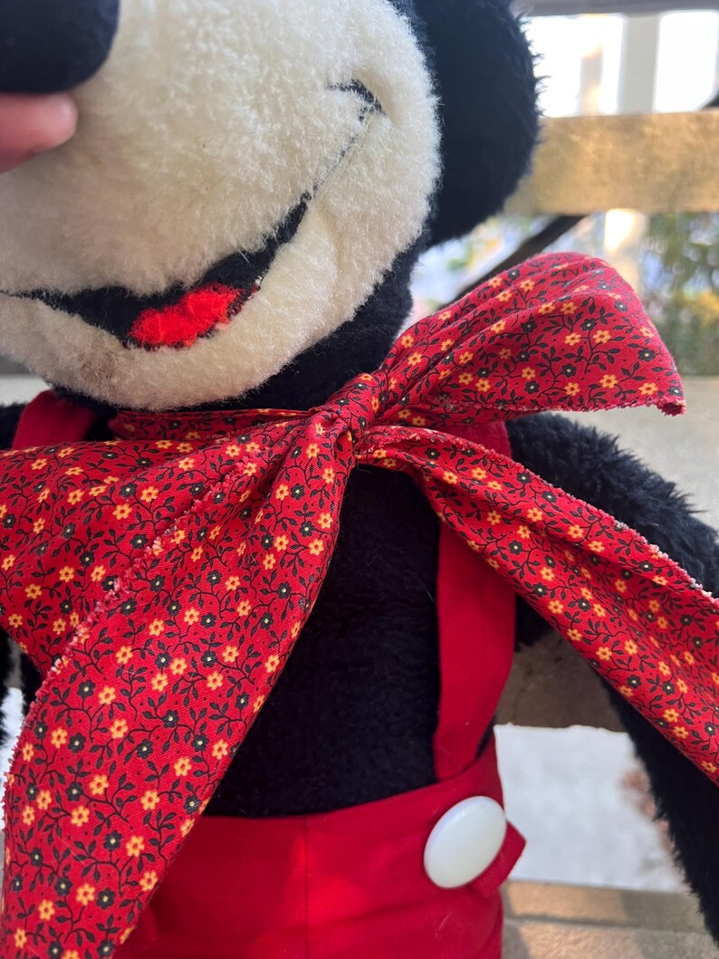 May include: A vintage plush toy featuring a black and white face with a red smile. The toy wears a red bow tie with a floral pattern and red overalls with a white button. The toy is a classic character.