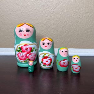 May include: A set of five traditional Russian nesting dolls, or matryoshka dolls. The dolls are painted in a green and white color scheme with pink flowers and black eyes. The largest doll is about 6 inches tall and the smallest doll is about 1 inch tall.