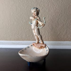 Puede incluir: Una pequeña figura vintage de Cupido, un querubín con arco y flecha, de pie sobre un plato en forma de concha. El plato es de color crema con detalles de estrellas azules y descansa sobre tres patas azules. La figura está pintada en tonos suaves y apagados.