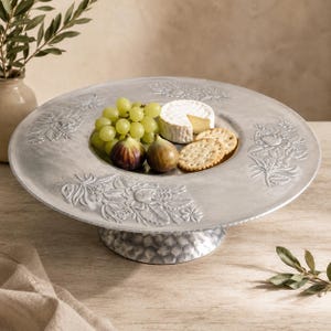 May include: A round silver-toned metal pedestal serving platter featuring an embossed floral pattern on the wide rim and a hammered texture on the base. The platter holds a cluster of green grapes, two fresh figs, a wedge of brie, and crackers.