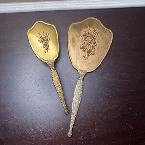Puede incluir: Un juego de dos espejos de mano dorados con diseños florales ornamentados en la parte delantera. Los espejos tienen un borde festoneado y un mango largo y detallado.