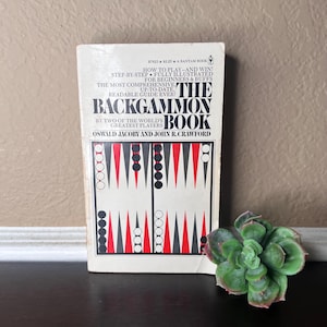 Può includere: Una copia vintage di "The Backgammon Book" di Oswald Jacoby e John R. Crawford. La copertina del libro è bianca con grafica nera e rossa e il testo recita "How to Play-and Win!" e "The Most Comprehensive, Up-to-Date, Readable Guide Ever!"