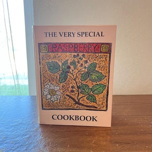 May include: A cookbook titled "THE VERY SPECIAL RASPBERRY COOKBOOK" with a light pink cover. The cover features a detailed illustration of a raspberry plant with green leaves, red berries, and a white daisy. The word "RASPBERRY" is in red letters.