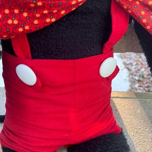 May include: A plush toy featuring a black body, red shorts with white buttons, and a red scarf with a floral pattern. The toy's design is reminiscent of a classic cartoon character.