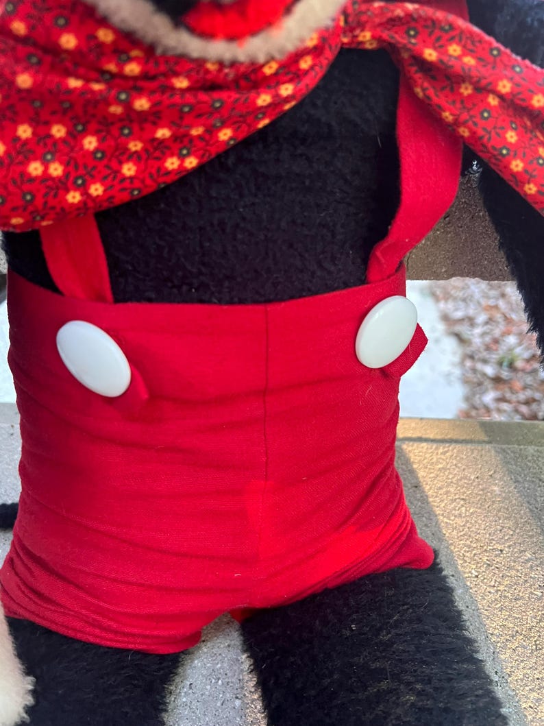 May include: A plush toy featuring a black body, red shorts with white buttons, and a red scarf with a floral pattern. The toy's design is reminiscent of a classic cartoon character.