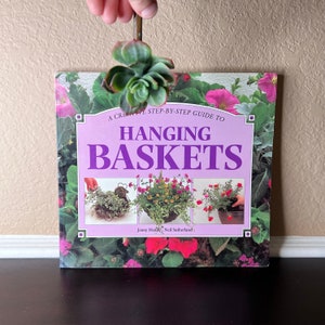 Puede incluir: Un libro morado titulado "Hanging Baskets: A Creative Step-by-Step Guide To Hanging Baskets" de Jenny Hendy y Neil Sutherland. La portada del libro presenta tres fotos de cestas colgantes con flores coloridas.