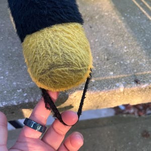 May include: A close-up of a plush toy's foot, featuring a black and yellow color scheme. The foot is yellow and fuzzy, with a black strap attached. The top portion of the toy is black and fuzzy.