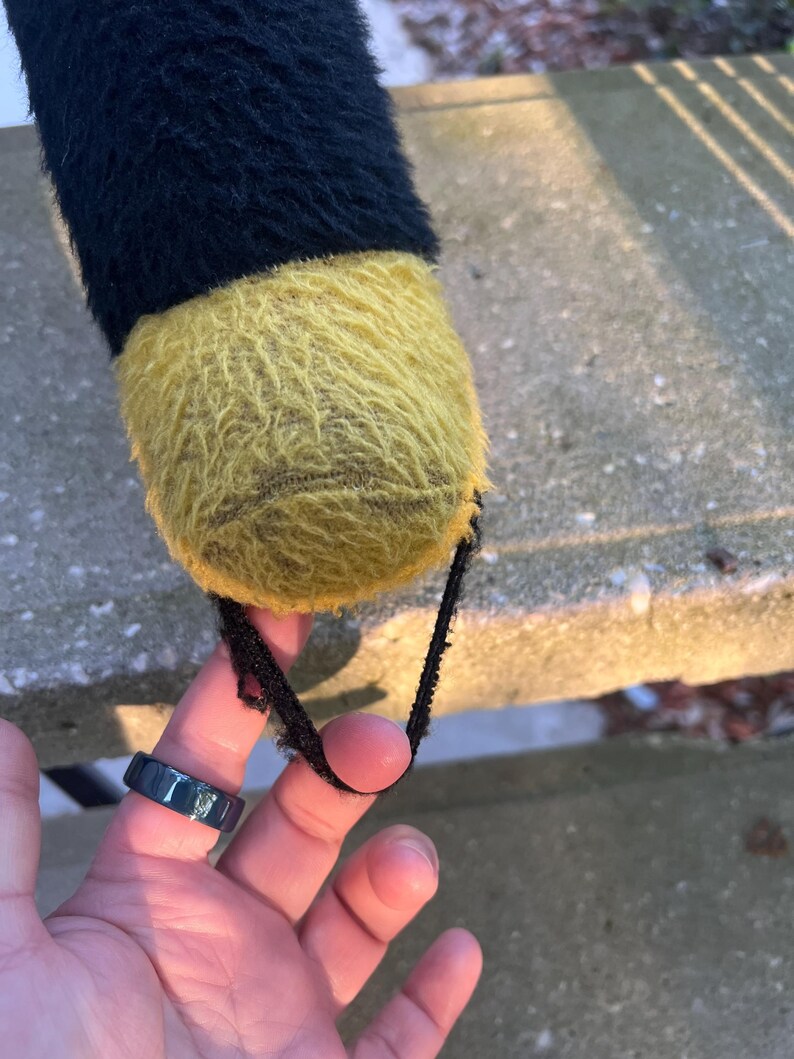 May include: A close-up of a plush toy's foot, featuring a black and yellow color scheme. The foot is yellow and fuzzy, with a black strap attached. The top portion of the toy is black and fuzzy.
