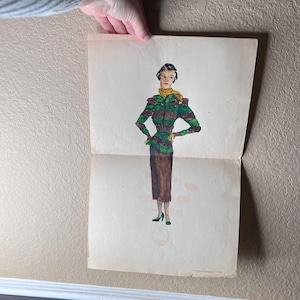 Puede incluir: Ilustración de moda vintage con una mujer con un atuendo verde, marrón y amarillo. El dibujo muestra una chaqueta de manga larga, una falda larga y una bufanda. La obra de arte está sobre papel envejecido, sostenido por una mano.