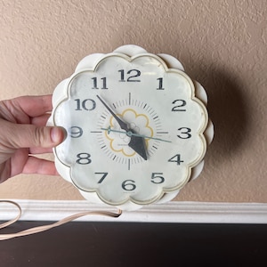 May include: A white, flower-shaped clock with black numbers and hands. The clock has a small, yellow flower design in the centre. The clock is plugged in and has a cord.