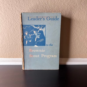 Può includere: Un libro blu con il titolo "Leader's Guide to the Brownie Scout Program". La copertina presenta un'illustrazione di tre ragazze in uniforme scout che giocano.