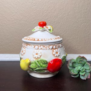 Peut inclure: Sucrier en céramique blanche avec un couvercle décoré de fruits rouges et jaunes, de feuilles vertes et de fleurs blanches. Le bol a une bordure brune et une base blanche.